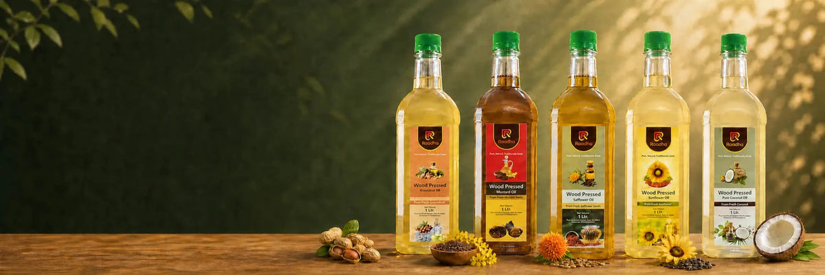 Five bottles of wood pressed oil arranged on a wooden table against a dark green leafy background. The bottles contain Groundnut, Mustard, Safflower, Sunflower and Coconut oil